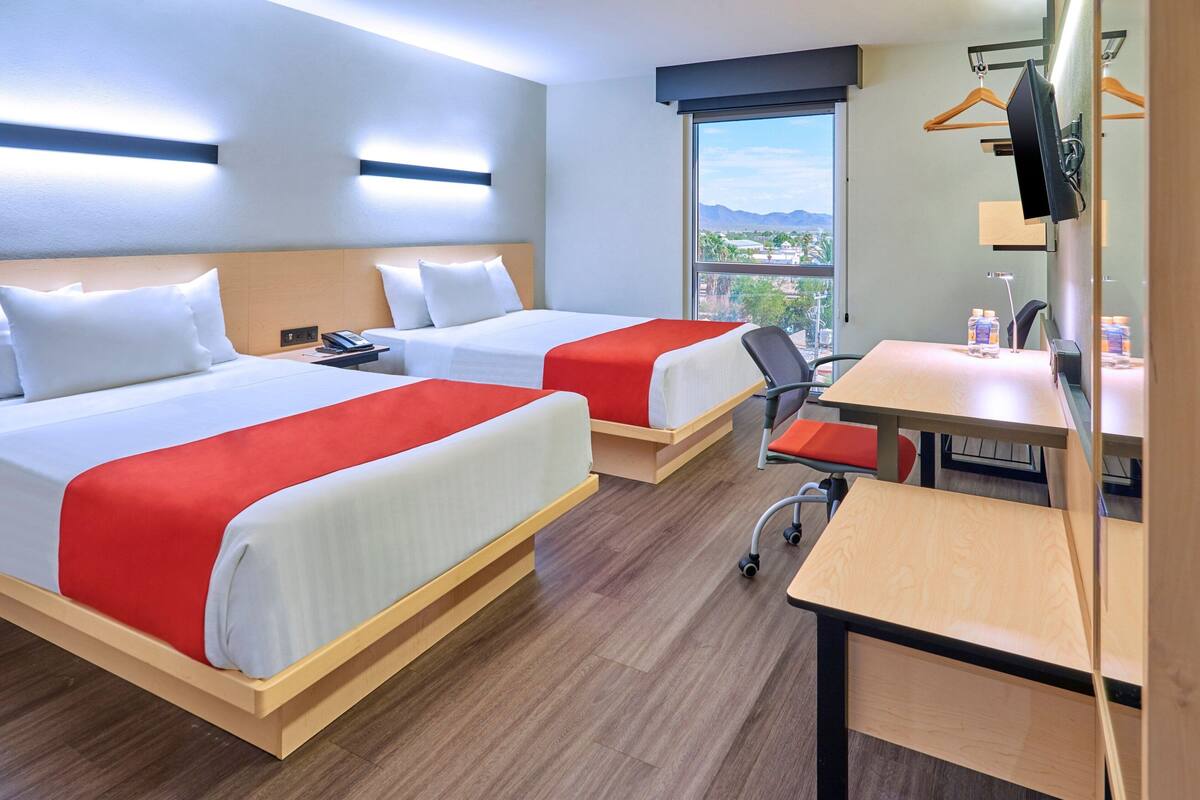 A hotel room with two beds, a desk, a chair, a TV, and a window with a view of mountains.