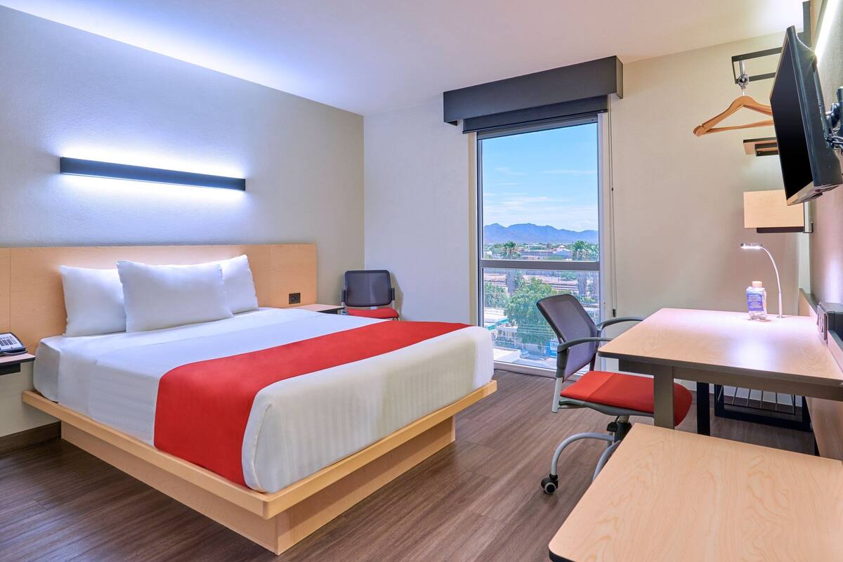 A hotel room with a bed, desk, chair, and a large window offering a city view.