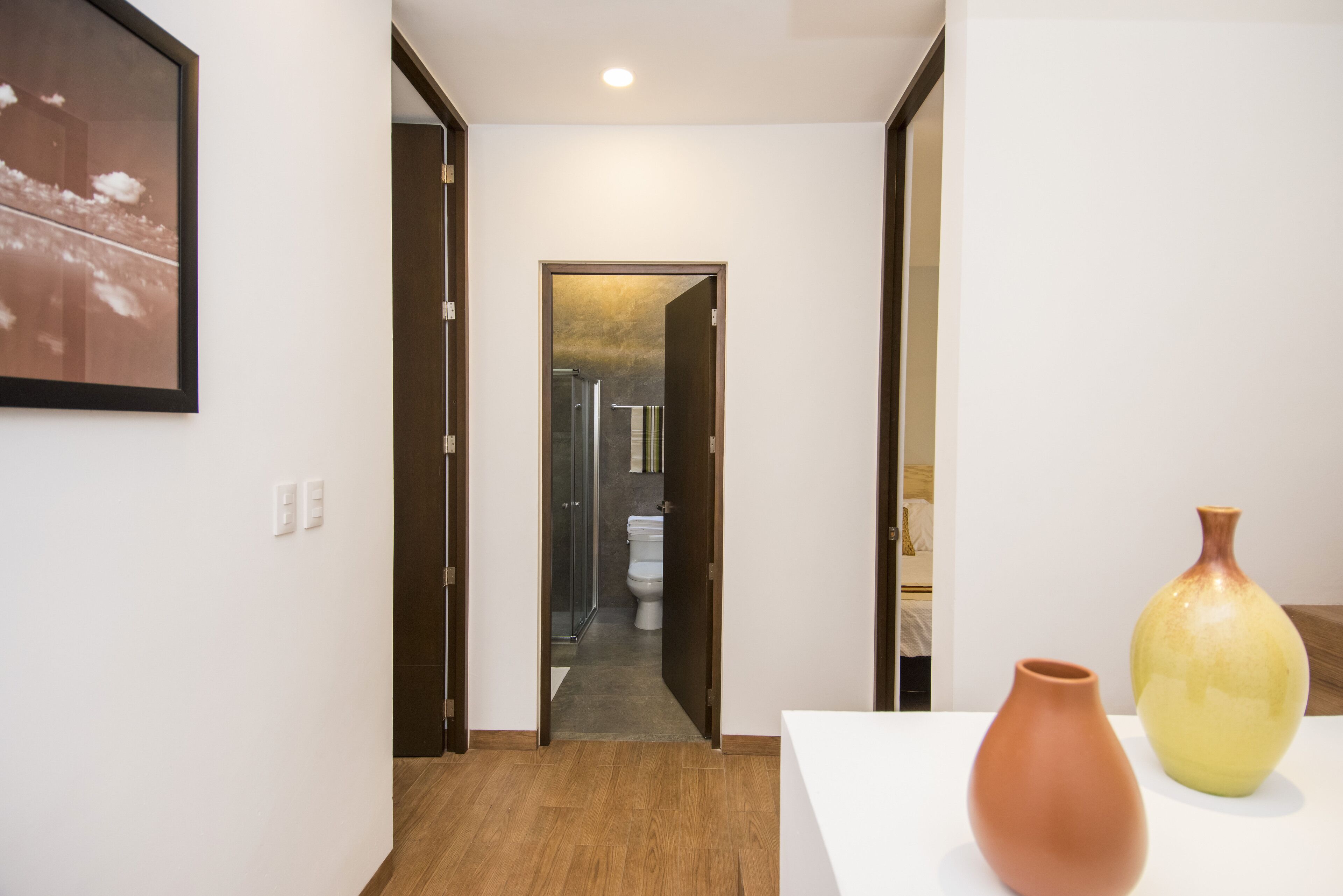 A hallway with a wooden floor, a white wall with a framed picture, and two vases on a table.