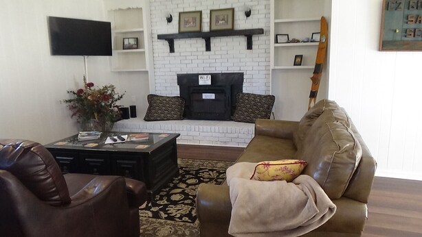 A living room with a white brick fireplace, a flat-screen TV mounted on the wall, and a brown leather sofa with a throw blanket. -  EZ - GO Lodging 3.7 mi Green Pond Landing(Lake Hartwell) Clemson Univ 15 mil. (Anderson)