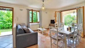 A modern living room with a grey sofa, a small white table, and a dining area with a white table and chairs. There are large windows providing natural light and views of greenery outside. - 2 198 / 5 000
Cozy cottage with private garden and terrace – peaceful setting (Le Bar-sur-Loup)