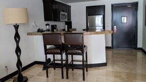 A modern kitchen with dark cabinetry, a stainless steel refrigerator, and a granite countertop. There are two bar stools and a floor lamp. - 2 Bedroom Serenity Pool View (San José del Cabo)