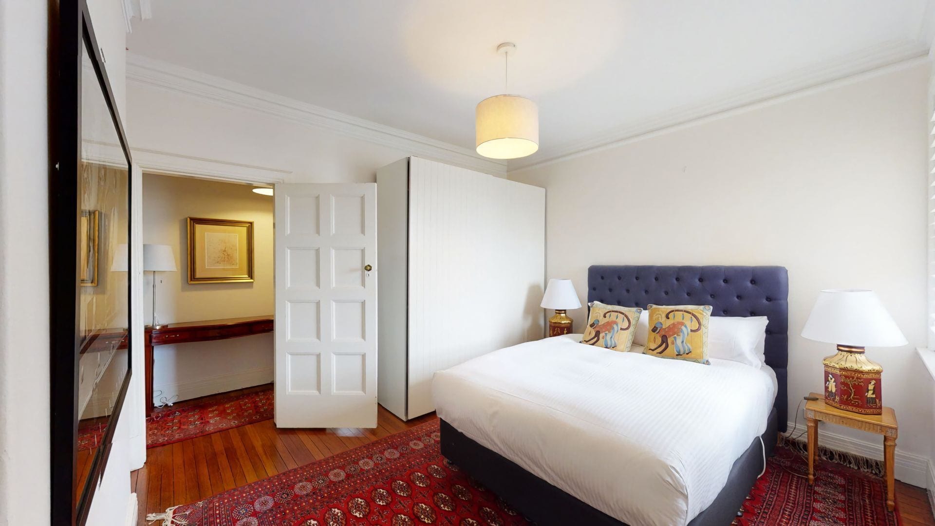 A hotel room with a large bed, two bedside tables with lamps, a red patterned rug, and a white door leading to another room.