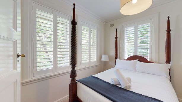 Un dormitorio con cama con dosel, paredes blancas y ventanas grandes con contraventanas.