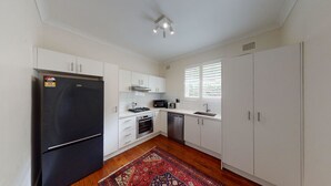Cocina compacta con armarios blancos, un refrigerador negro, un microondas, una estufa y un lavavajillas. Hay una ventana con persianas y una alfombra estampada sobre el suelo de madera.