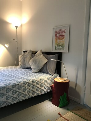 Een slaapkamer met een bed, kussens, een nachtkastje, een lamp, een lijst met een poster en een rode stronk.