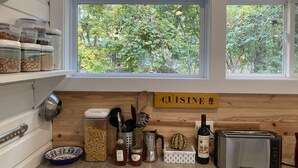 Una cocina con vista a árboles por la ventana, un cartel que dice 'CUISINE' y diversos utensilios de cocina, como pasta, miel, frascos y un tostador.
