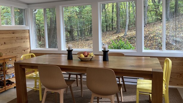Comedor con mesa de madera, cuatro sillas y vistas a un bosque a través de amplios ventanales.