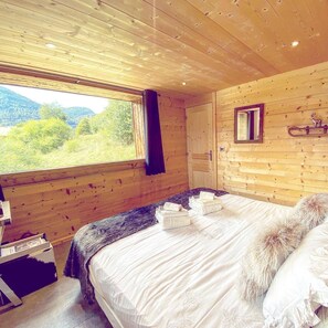 Un dormitorio tipo cabaña de madera con un ventanal grande, una cama con sábanas blancas y un espejo en la pared.