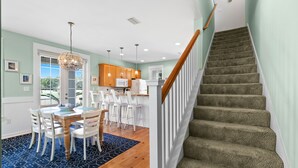 Una cocina moderna con armarios de madera, una zona de comedor con sillas blancas y una escalera con barandilla de madera.