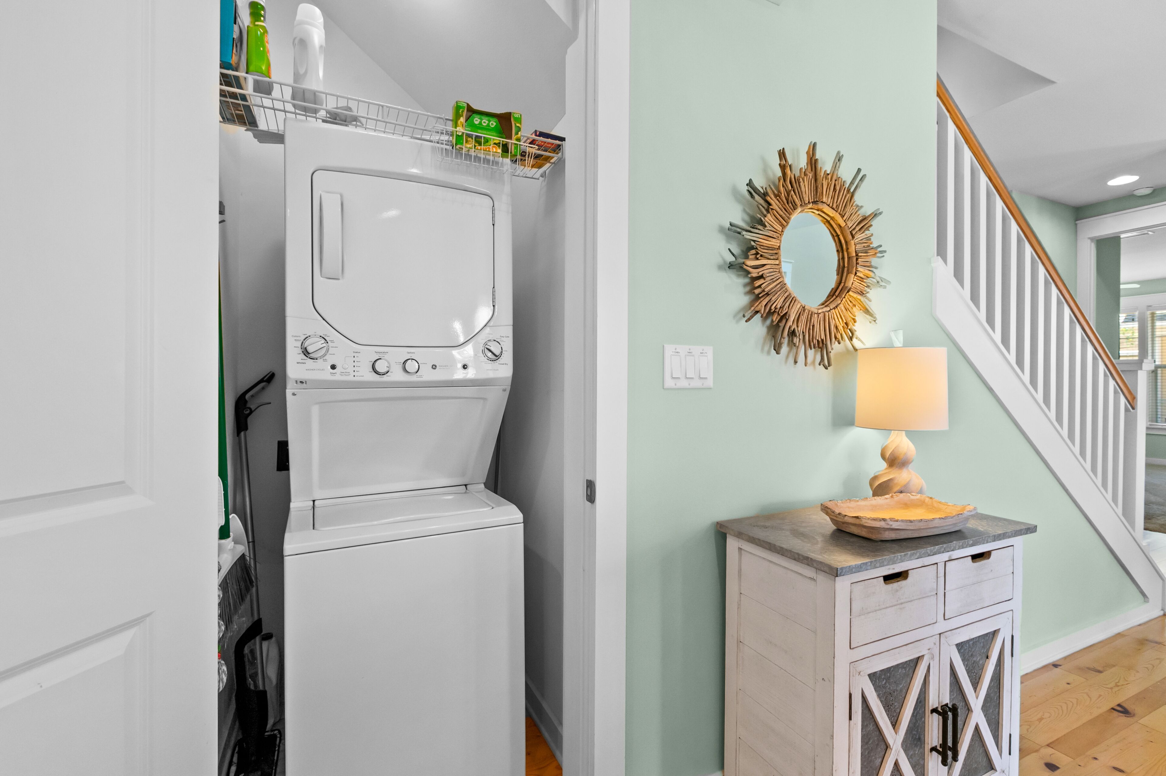 Une buanderie équipée d’un lave-linge et d’un sèche-linge superposés, d’un meuble en bois avec des tiroirs, d’un miroir décoratif et d’un escalier.
