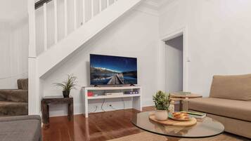 A modern living room with a staircase, a flat-screen TV on a white entertainment unit, a round glass coffee table, and a beige sofa.