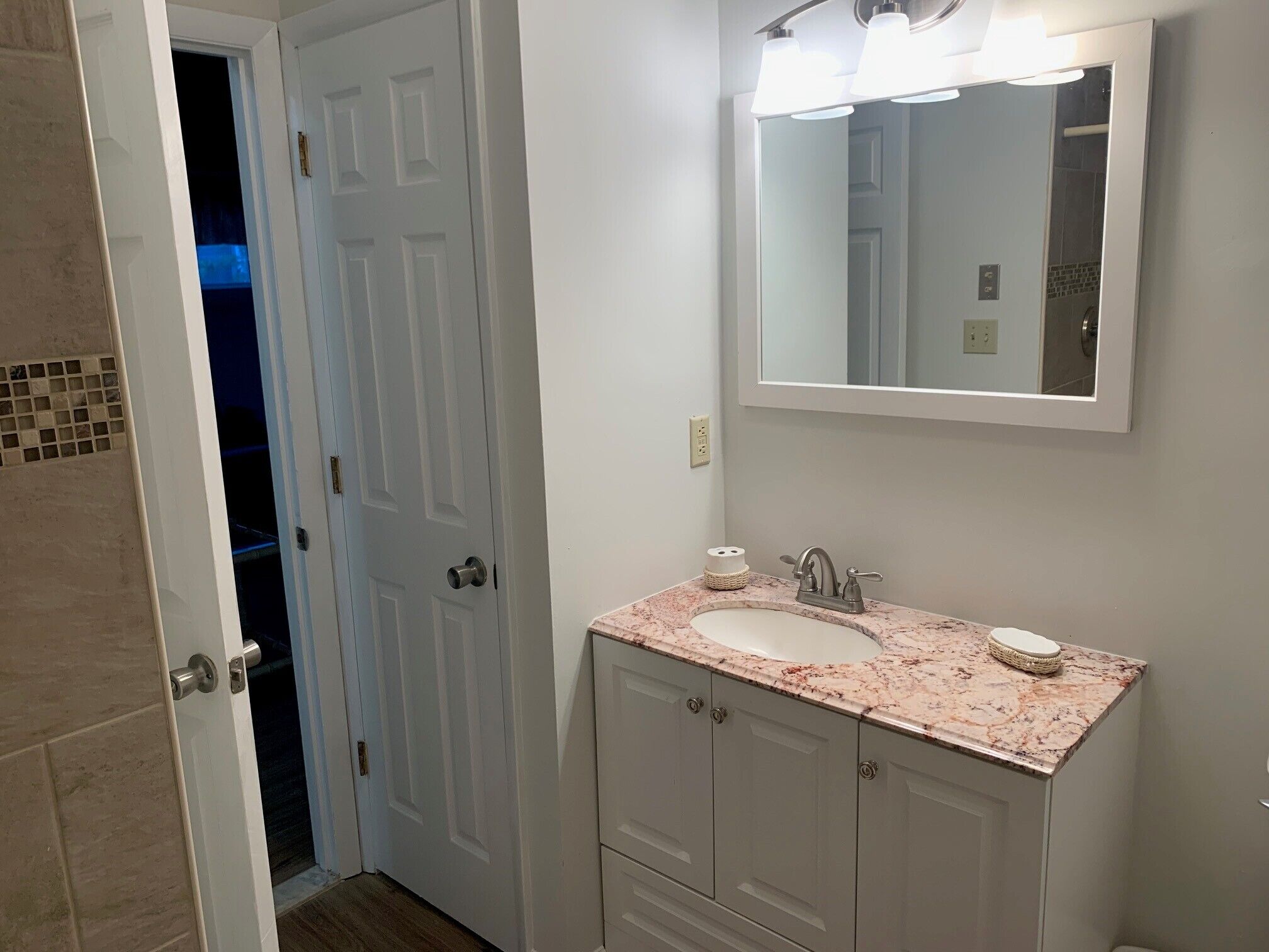 A bathroom with a white door, a sink with a marble countertop, a mirror above the sink, and a small basket on the countertop.