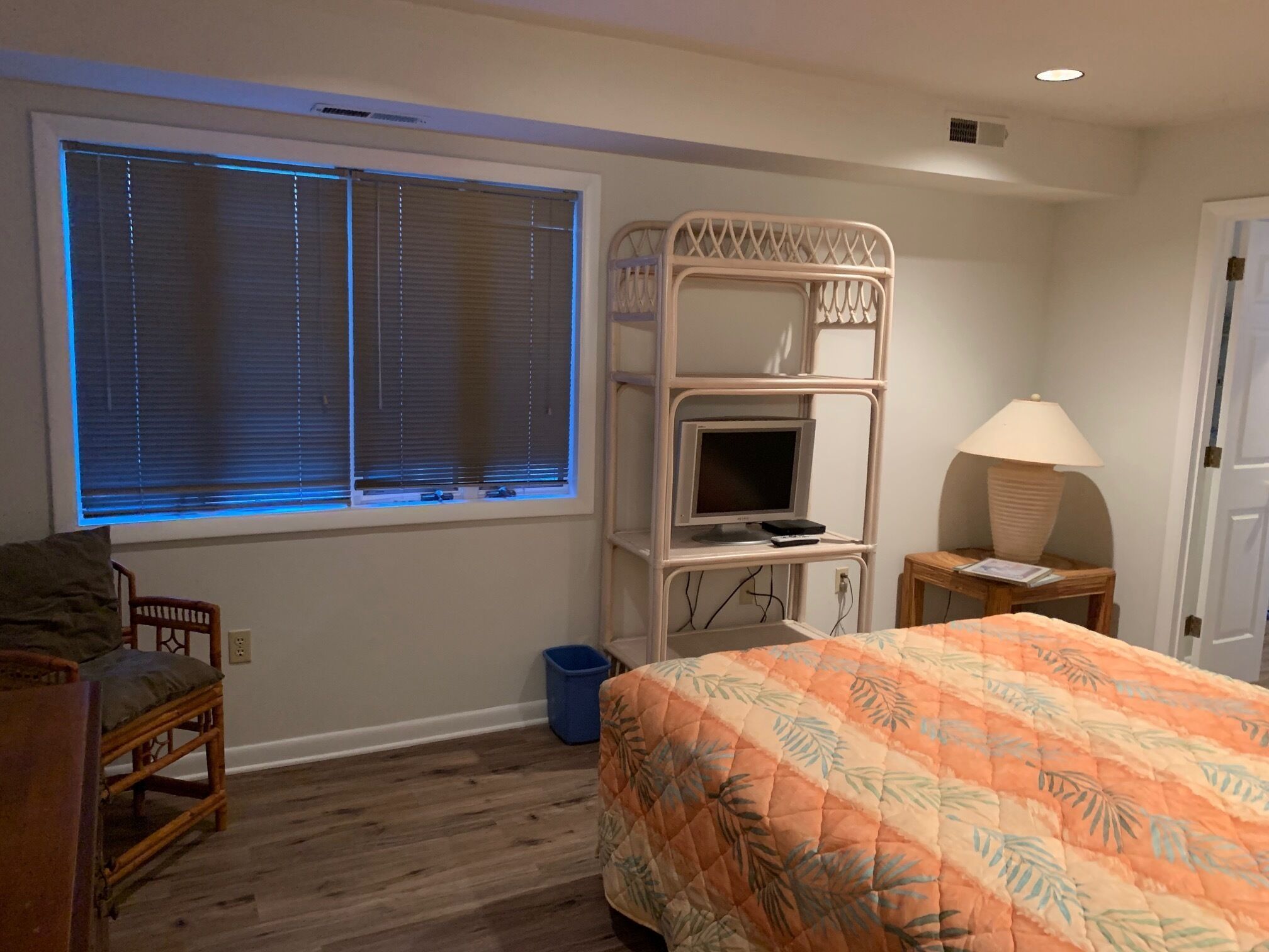 Une chambre à coucher avec un lit, un téléviseur posé sur une étagère, une lampe, une chaise et une fenêtre avec des stores.