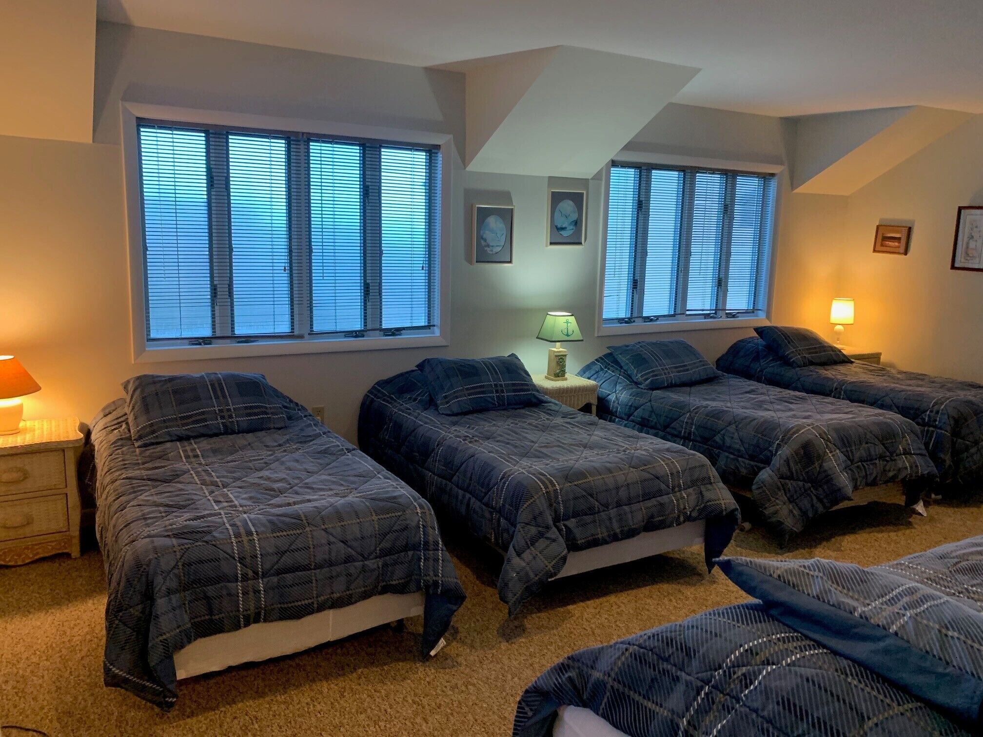 A bedroom with three beds, large windows with blinds, a nightstand with a lamp, and framed pictures on the wall.