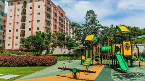 A playground with a seesaw, slides, and climbing structures, surrounded by a well-maintained garden and a multi-story residential building in the background. - Chateau Elysee - Seine Cluster (Parañaque)