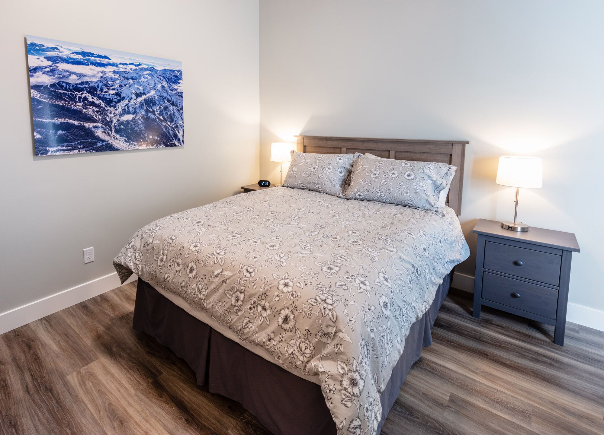 A bedroom with a bed, two bedside tables, a nightstand, a wall-mounted lamp, and a framed picture.