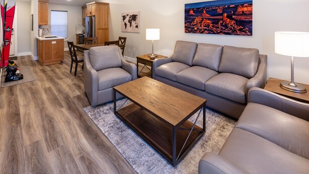 Ein modernes Wohnzimmer mit einem grauen Sofa, einem Holztisch und einem Essbereich mit Holzschränken und einem Kühlschrank.