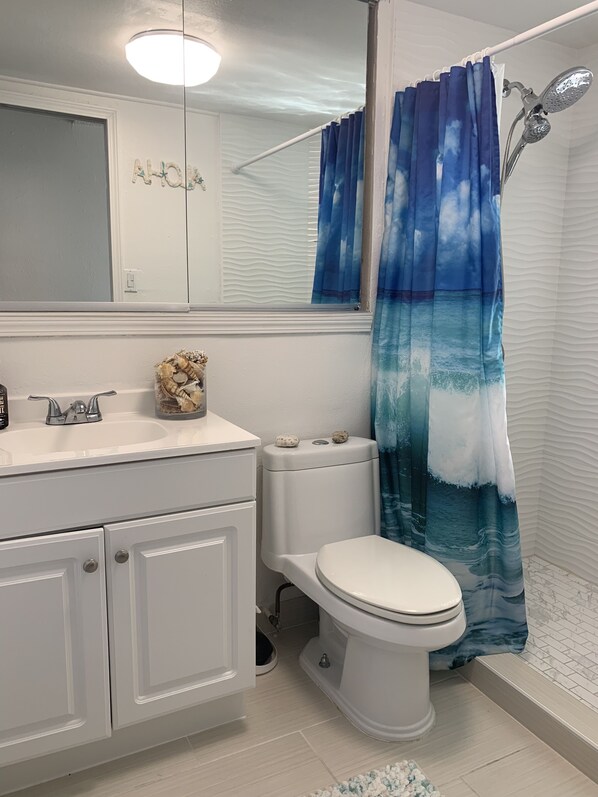Une salle de bain avec des toilettes blanches, un lavabo surmonté d’un miroir et un rideau de douche orné d’un motif représentant l’océan.
