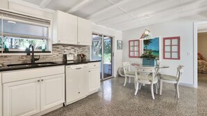 A modern kitchen with white cabinets, a black countertop, and a dining area with a glass table and white chairs. - Old Florida Key West Cottage w/ river access (Jensen Beach)