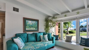 A teal sofa with patterned cushions, a framed painting of an underwater scene, and a large window with a view of greenery and a parked car. - Old Florida Key West Cottage w/ river access (Jensen Beach)