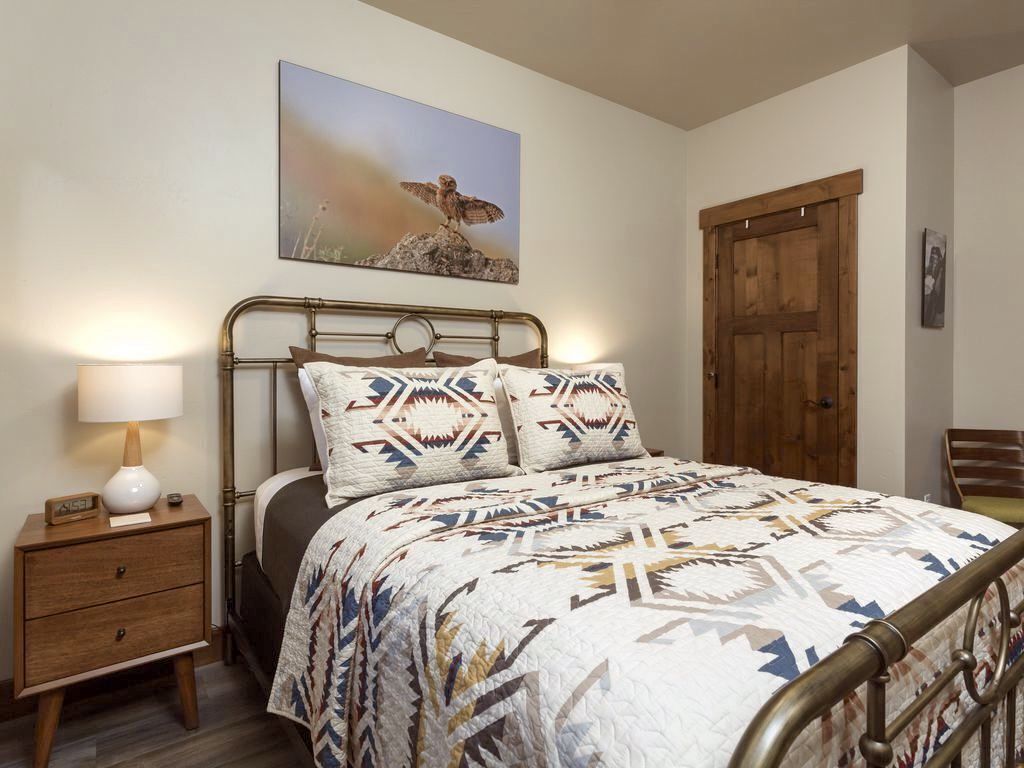 Une chambre à coucher avec un lit, une table de chevet, une lampe, une porte en bois et un tableau accroché au mur.