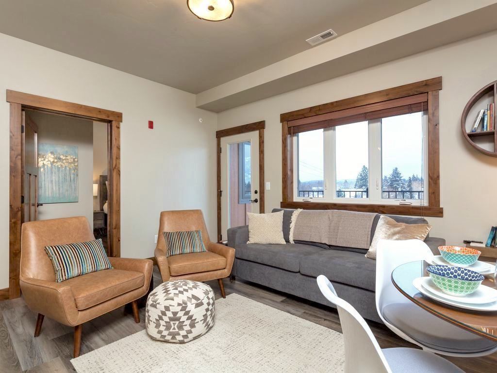 A living room with a grey sofa, two armchairs, a round coffee table, and a dining table with chairs. There are large windows with a view of trees and buildings.