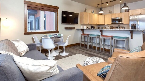 A modern living room with a grey sofa, white chairs, and a kitchen area with wooden cabinets, a microwave, and a refrigerator. - Beautiful 3-bed/2-bath Condo Looking Into Glacier National Park! (Columbia Falls)