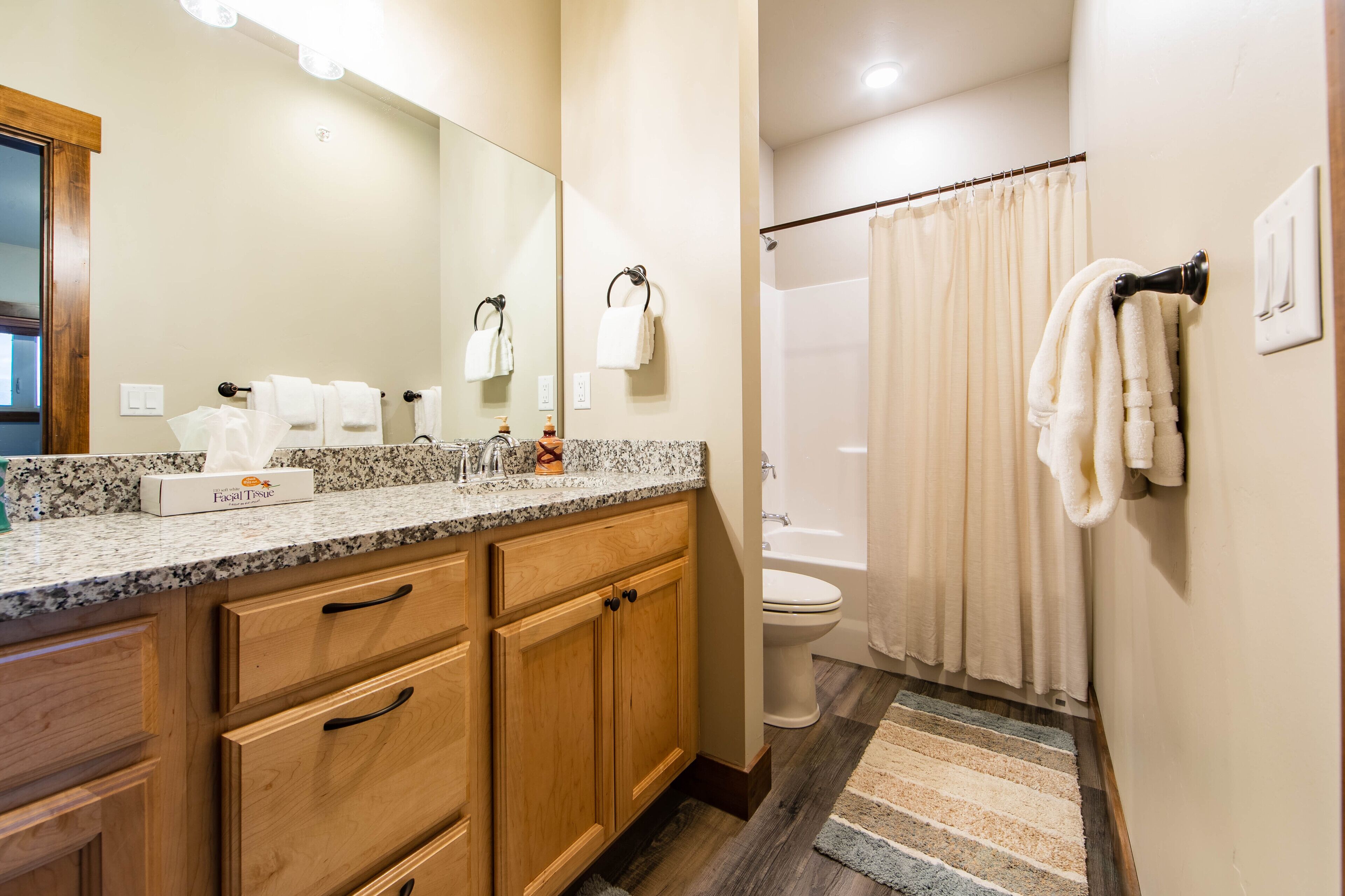 Une salle de bain avec un comptoir en granit, des armoires en bois, des toilettes blanches, une douche avec un rideau beige et un miroir avec éclairage intégré.