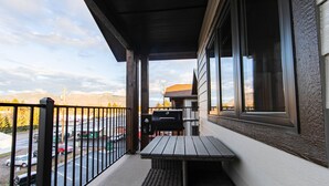 A balcony with a table and chairs, a grill, and a view of a parking lot and mountains. - Beautiful 3-bed/2-bath Condo Looking Into Glacier National Park! (Columbia Falls)