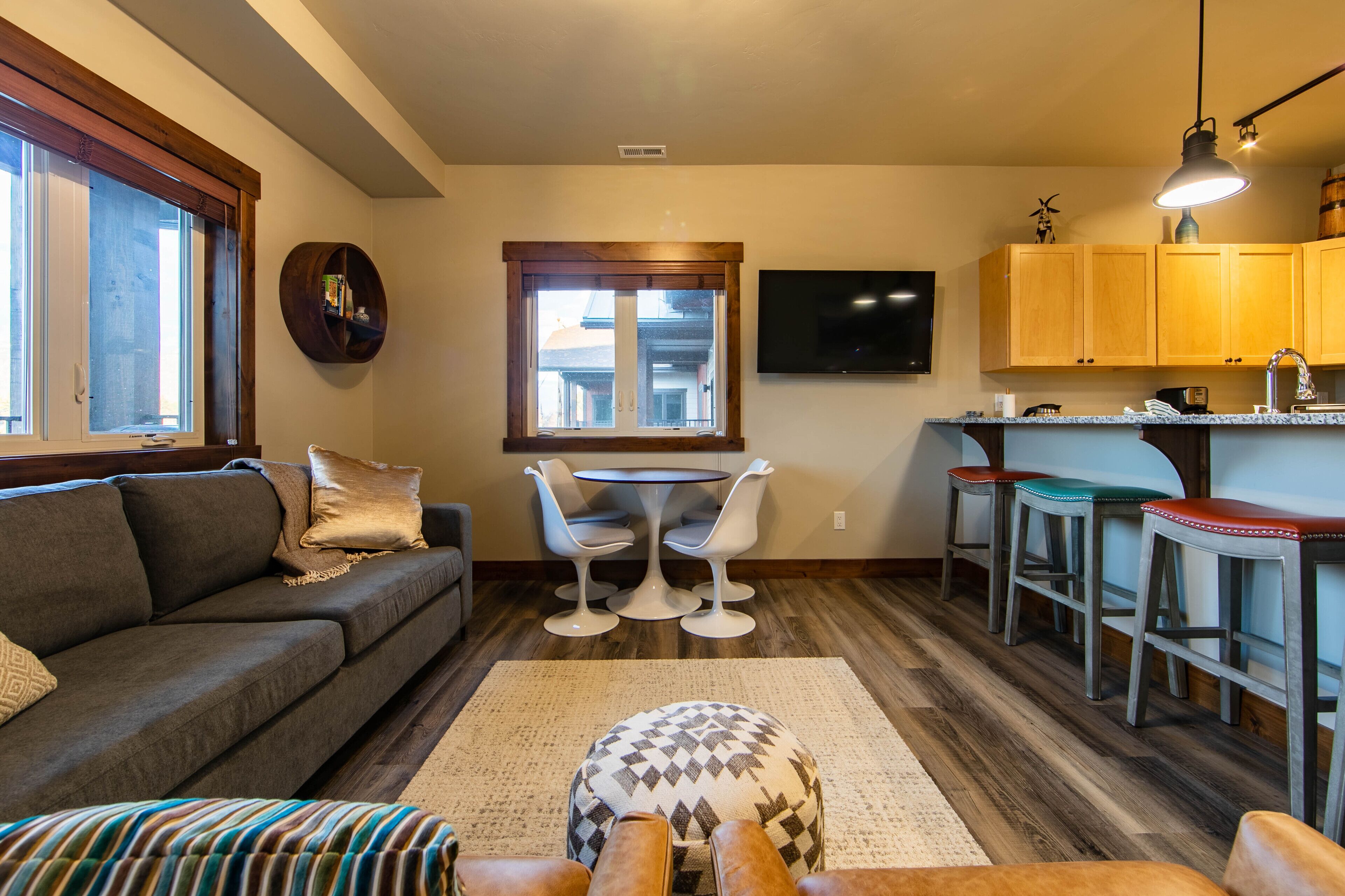 A living room with a sofa, a coffee table, and a patterned ottoman. There is a dining area with a round table and chairs, and a kitchen area with wooden cabinets and a bar stool.