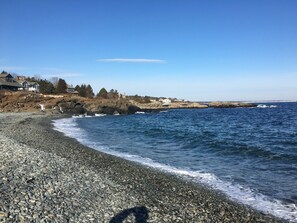 Une côte rocheuse où les vagues s’écrasent sur les galets, un ciel bleu dégagé et des maisons en arrière-plan.