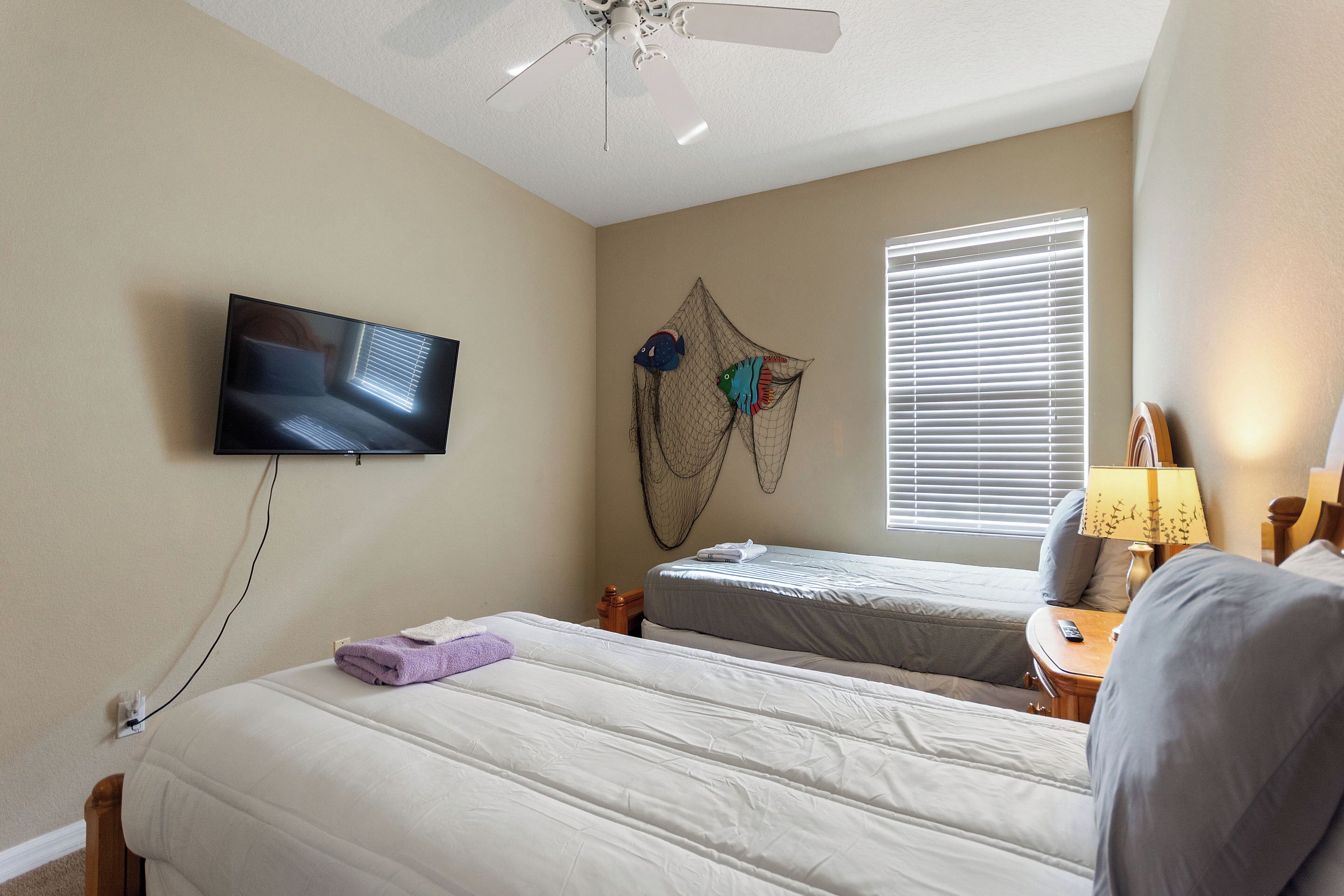 A hotel room with two beds, a ceiling fan, a TV mounted on the wall, and a window with blinds.