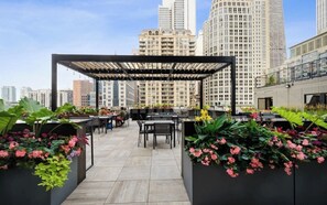 An outdoor rooftop terrace with tables and chairs, surrounded by planters with flowers, and a view of tall buildings in the background. - Luxury 1BR Condo – Views, Comfort, & Style (Chicago)