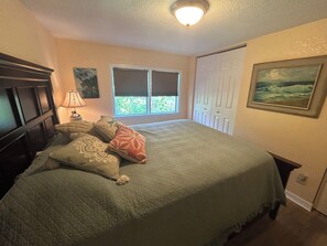 A bedroom with a bed, a nightstand, a window with blinds, a door, and a painting on the wall.