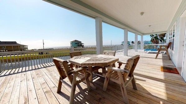 A wooden deck with a table and chairs, overlooking a beach with a white fence and a small building.