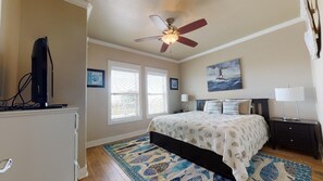 A bedroom with a bed, a ceiling fan, a TV, a desk, and a window.
