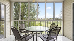 En täckt uteplats med ett glasbord och fyra stolar, stora fönster och en takfläkt.