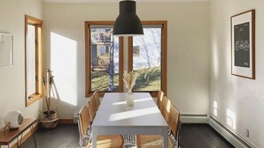 Un comedor con una mesa blanca, sillas de madera, una lámpara colgante negra y un ventanal con vistas a un edificio y árboles.