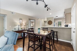 Un coin repas avec une table à plateau en verre et des chaises en bois, une cuisine avec une horloge et un couloir avec une porte.