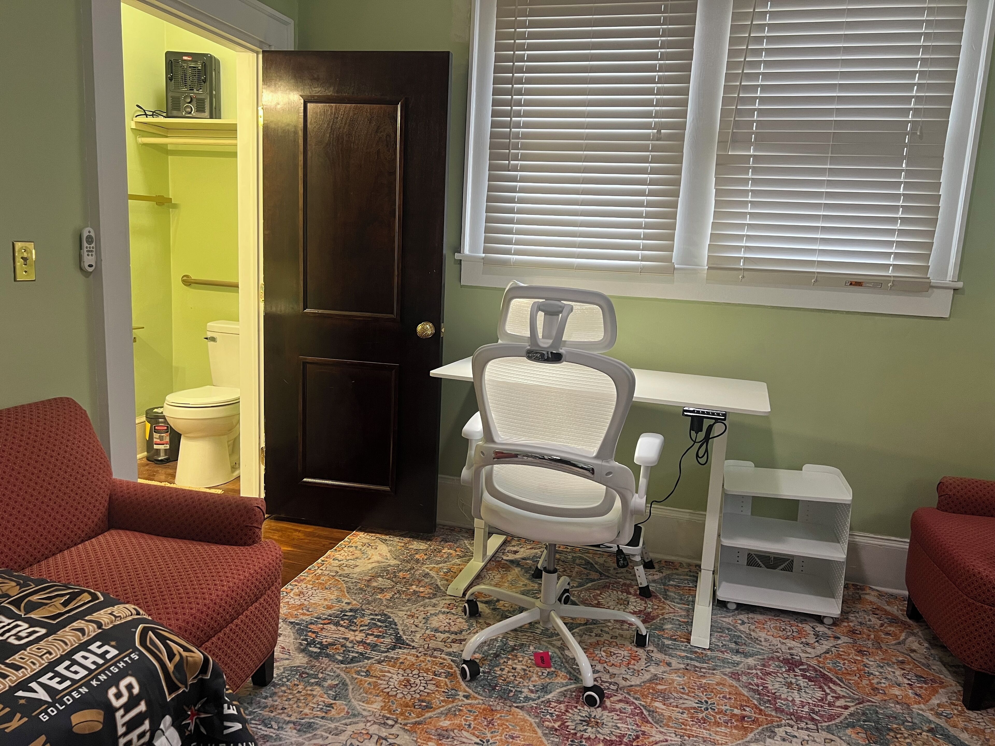 Une petite pièce bien éclairée, avec un bureau, une chaise et deux fauteuils rouges. Une porte donne sur une salle de bain, et une fenêtre avec des stores se trouve sur le mur.