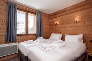 Une pièce aux murs lambrés, comprenant un lit, deux fenêtres avec des rideaux, un radiateur fixé au mur, et offrant une vue sur un paysage enneigé.