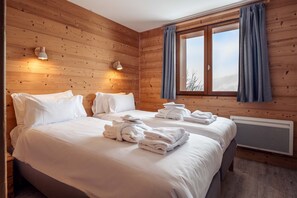 A hotel room with wooden walls, a bed with white linens, two bedside lamps, a window with curtains, and a radiator.