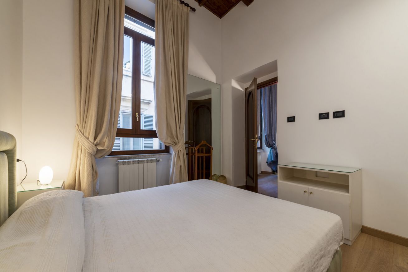 A bedroom with a bed, a bedside lamp, a window with curtains, a mirror, a dresser, and a door leading to another room.