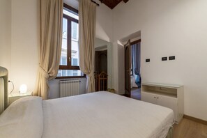 Quarto com cama, luminária de cabeceira, janela com cortinas, espelho, cômoda e porta para outro espaço.