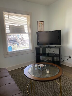 A living room with a television on a stand, a glass coffee table, a sofa, and a window with blinds.