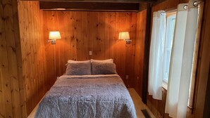 Quarto com painéis de madeira, cama, duas luminárias fixadas na parede e janela com cortinas.