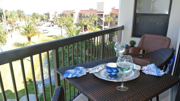 A balcony with wicker furniture, a table with a glass of water, and a view of a residential area with palm trees and buildings. - Ocean Village Club I33, Elevator, 2 Pools (1 Heated) Ocean View (St. Augustine)