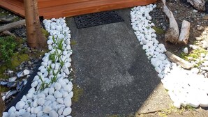 Ein Haus mit einer Holztür, einer Treppe aus Holz, die zu einer Veranda führt, und einem kleinen Garten mit weißen Kieselsteinen und einem Baum.