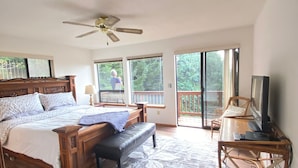 Ein Schlafzimmer mit einem großen Bett, einer Bank, einem Fernseher und einem Balkon mit Blick auf Bäume.
